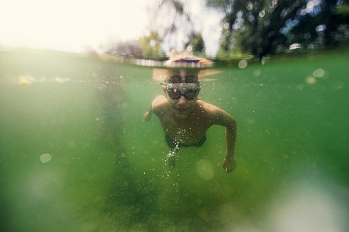 Teenage boy enjoying swimming underwater in a small pond or lake in the Austrian countryside. The boy is swimming and smiling at the camera.\nCanon R5