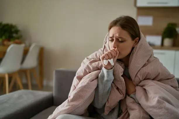 Photo of Caucasian young woman coughing and sitting under the duvet at home
