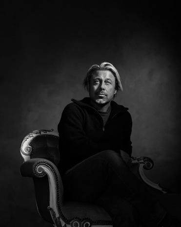 Man with blond hair and a stubble beard in black woolen sweater sits on a vintage bench. Black and white photo.