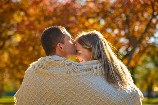 Beautiful couple man woman in love. Man and woman wrapped in beige blanket hugging together in park on autumn fall sunny day. Authentic real people.