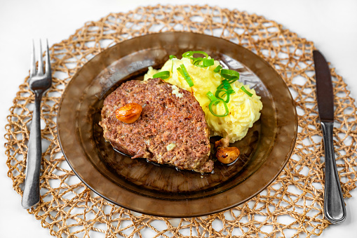 Baked minced beef, garlic and mashed potato on plate, cutlery and bamboo pad on table. Serving size, lunch or dinner.
