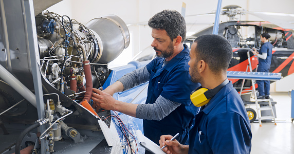 Male mechanics with multimeter and clipboard checking helicopter engine in airplane hangar.