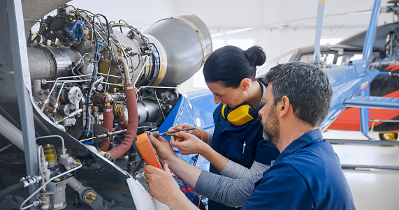Male and female mechanics with multimeter working on helicopter engine in hangar.