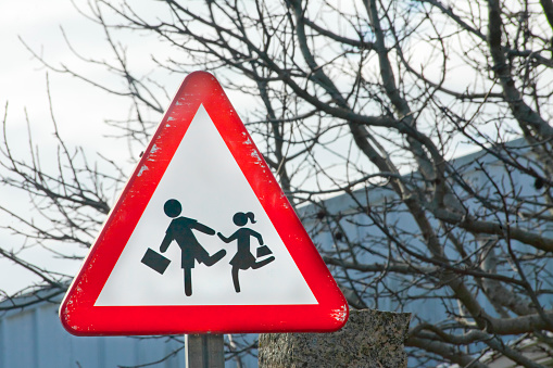 Triangle warning pedestrians road sign, children,school building nearby. Galicia, Spain. Copy space on the right side of the image, bare tree and mountainous landscape in the background.