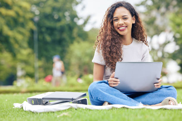 한 젊은 히스패닉 여성이 열린 들판에 밖에 앉아있는 동안 노트북으로 작업하고 있습니다. 대학 캠퍼스에서 온라인으로 공부하기 위해 컴퓨터를 사용하는 동안 웃고있는 아름다운 혼합 인종 여 - laptop computer grass nature 뉴스 사진 이미지