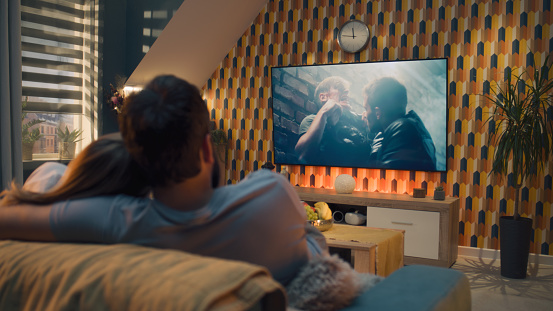 Couple on sofa in living room, watching action movie on TV or criminal blockbuster on streaming service, talking and discussing acting, resting at home on weekend. Home theater in modern apartment.