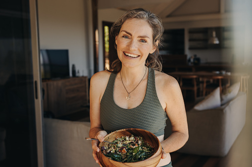 Cheerful vegan woman smiling at the camera while holding a buddha bowl. Mature woman enjoying a plant-based breakfast after working out at home. Happy senior woman taking care of her ageing body.