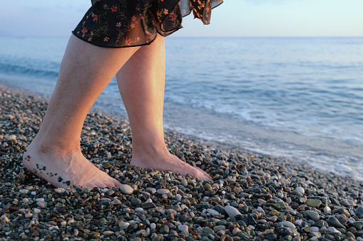 Women's feet at the water's edge on a pebble beach. Woman standing by the sea in the sun. The concept of rest on the sea, unity with nature, travel