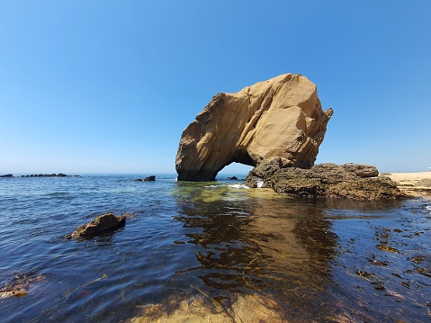 A large stony formation with an arch on the water against a blue sky