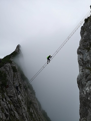 A vertical shot of a climber crossing from one cliff to another over a rope bridge in a foggy weather