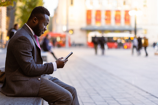 Black businessman reading text message. He is sitting  outdoors in a city square, taking a break