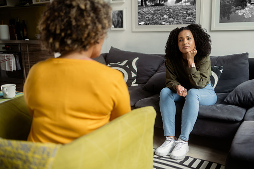 An African-American teen girl sits on a couch and talks to the unrecognizable female psychotherapist at her office. The girl rests her head on the hand and looks like she's uncomfortable.