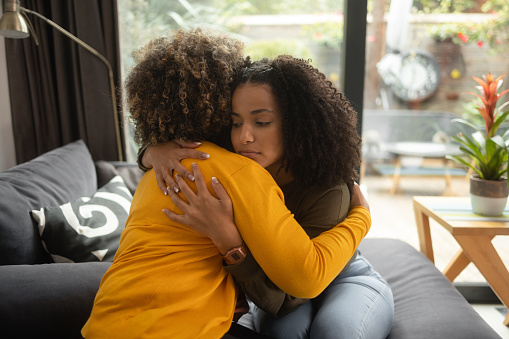 An African-American female psychotherapist comforts the teenage female patient by hugging her. Both of them have the afro hairstyle.