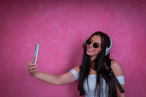 A portrait of a young stylish woman with sunglasses and headphones. She holds a smartphone and takes selfies in a photographic studio.