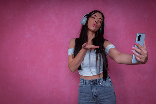 A portrait of a young stylish woman with headphones, taking selfies and sending a kiss in a photographic studio.