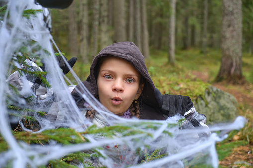 a child in a halloween costume makes faces, a girl hides behind a tree decorated for halloween, a halloween party in the forest, a tree in a cobweb and halloween decorations