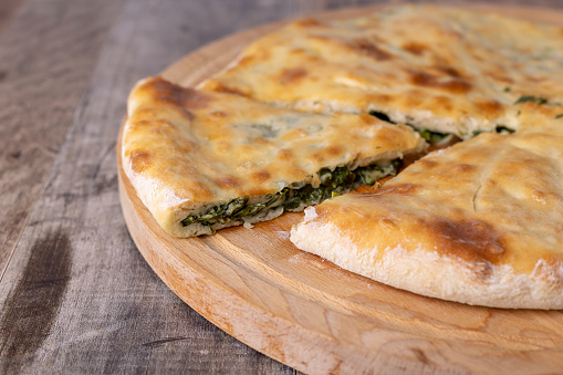 Pie with spinach, herbs and Suluguni cheese on a wooden board. The whole cake with the piece cut off. Traditional round flat cake with filling. Homemade pastries of Caucasian cuisine. Selective focus, close-up.