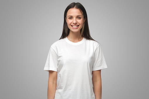 Portrait of smiling young woman in white t-shirt looking at camera, isolated on gray background