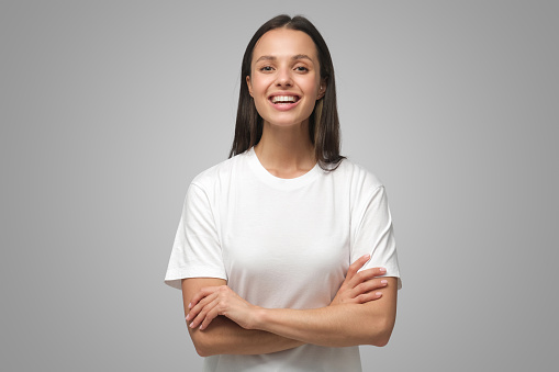 Woman crossed arms. Portrait of young smiling female standing in white t-shirt, isolated on gray background