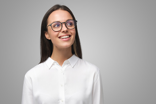 Young female in white shirt and eyeglasses looking aside with dreamful expression waiting for something good to happen, isolated on gray