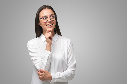Young business woman looking aside through big spectacles dressed in formal clothes dreaming or thinking of new ideas, isolated on gray, copy space on right side