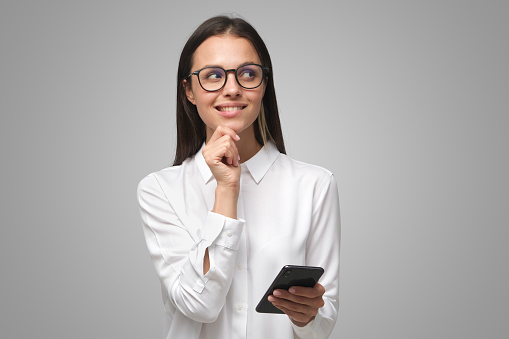 Cunning woman making plans while holding cellphone in hand and looking to right side through big glasses with smile, isolated on gray background with copy space