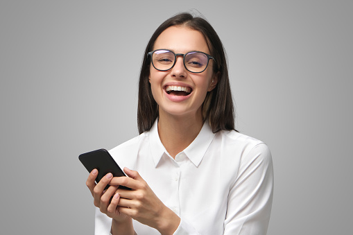 Young business woman laughing while reading text messages from colleagues on her phone, dressed in white collar shirt, isolated on gray background