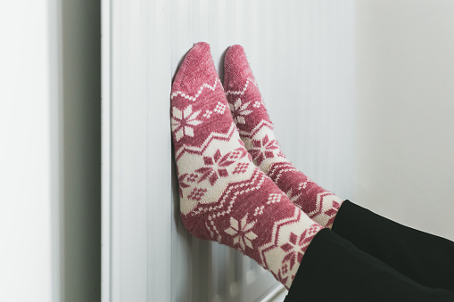 Color image depicting a woman warming her feet on the radiator while wearing festive fluffy winter socks.