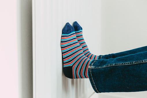 Color image depicting a man warming his feet on the radiator while wearing stripy socks.