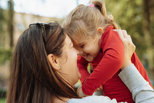 Mid adult mother and little girl playing outdoors on sunny autumn day in park. Cheerful girl wearing red shirt.