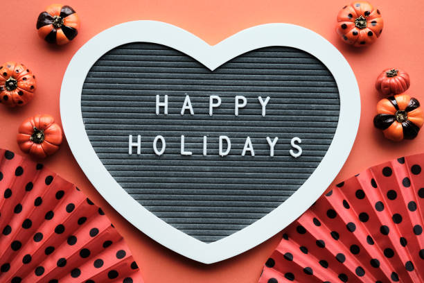 Halloween orange black flat lay background with decorations. Heart shaped felt letter board with text Happy Holidays. Orange fans with polka dots, painted mini deco pumpkins with black stripes and circles.