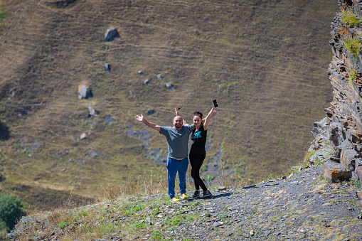 A man and a girl in the mountains look at the camera with open arms