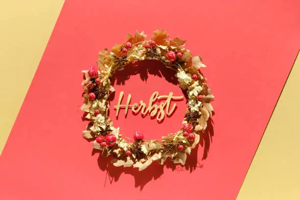 Dried floral wreath made of dry Autumn leaves and berries. Word Herbst means Autumn in German language. Seasonal Fall flat lay on red and yellow paper background. Direct sunlight with shadows.