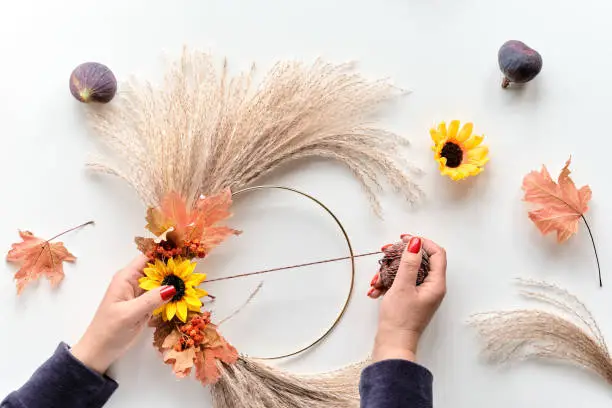 Hands making dried floral wreath from dry pampas grass and Autumn leaves. Hands tie decorations to metal frame with cord. Flat lay on white table, soft natural sunlight.