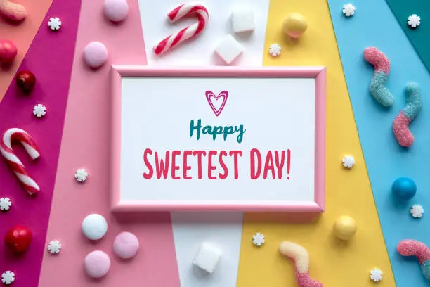 Text Happy Sweetest day in pink frame. Assorted sweets, chocolates, confectionery on multicolor layered paper. Flat lay, top view. Sweetest day holiday bright colorful greeting design.