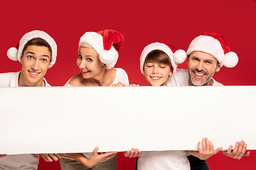 A wonderful happy family wearing Santa Claus hats and smiling to the camera- two sons, mom and dad. Friendly people pose in the studio, holding a white empty board in their hands. Red background. Real people lifestyle. Xmas time.