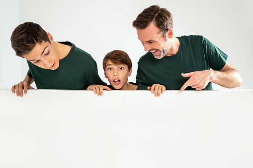 Dad and his two sons pointing on an empty white board, smiling. Studio shot. Real people emotions. Sale concept.