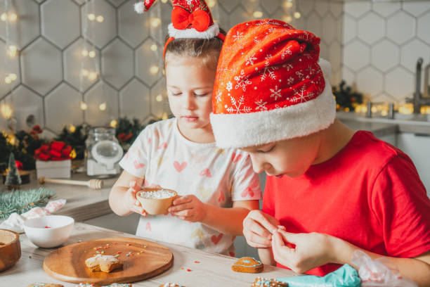 Girl 3 years old and boy 8 years old in Santa hat decorate gingerbread cookies with icing. Siblings in Christmas kitchen stock photo