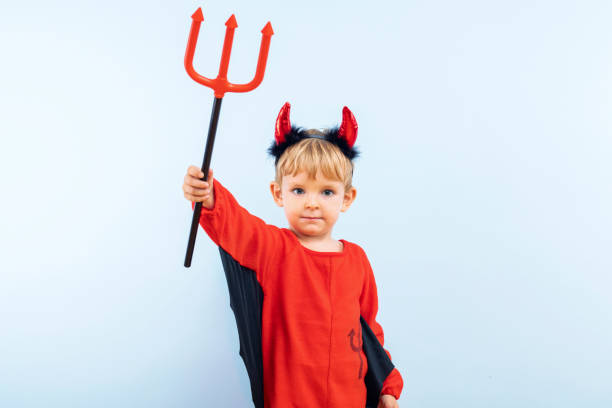 Happy Halloween. Cute little boy in devil halloween costume with horns and trident on light blue background.