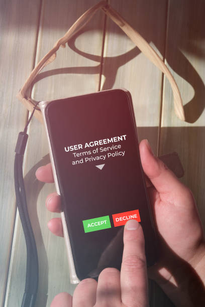 istock Finger press Refuse button on mobile phone screen. User disagree with Terms of Service and the Privacy Policy mobile app. Close-up on device with hands on wooden table with reading glasses. 1433415112