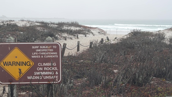 Ocean beach sandy dunes, Monterey, California misty coast, USA. Foggy rainy autumn, winter weather, grey cloudy sky. Trail path on shore near cold sea waves. Danger sign, life threatening rip currents