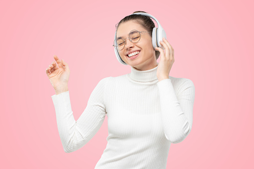 Young female in eyewear and headphones, dancing to sounds of music with eyes closed, feeling happy and smiling, isolated on pink background