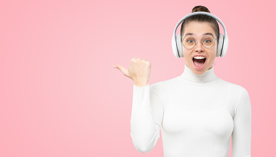 WOW! Horizontal banner of young shocked girl listening to music in wireless headphones, showing to copy space with excited face and open mouth, isolated on pink background
