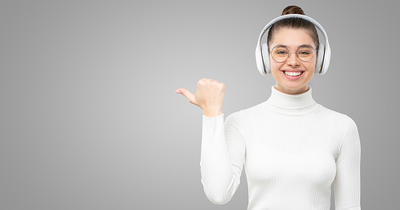 Horiozntal shot of girl in headphones, dressed in white turtleneck sweater, pointing at copy space on left, smiling happily, isolated on gray background