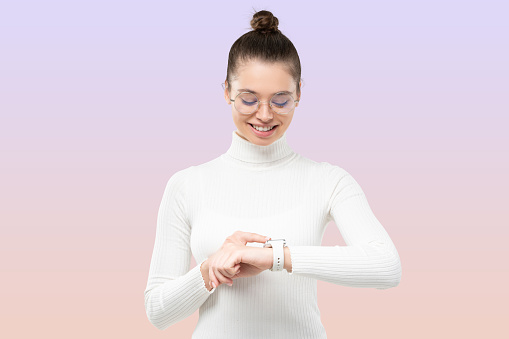 Young female in white sweater and glasses looking at smartwatch on her wrist, checking time, glad she is not late, isolated on purple gradient background