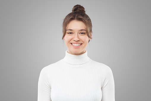 Portrait of beautiful young woman isolated on gray background, feeling positive and shy, wearing white turtleneck sweater, hair tied in bun with loose strands