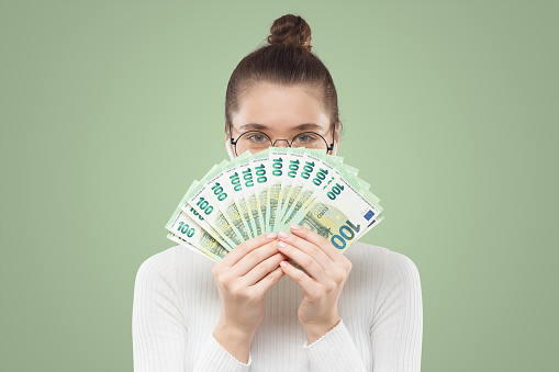 Young student girl in glasses and turtleneck hiding behind fan of 100 hundred euro banknotes, satisfied with her lottery prize, isolated on green background