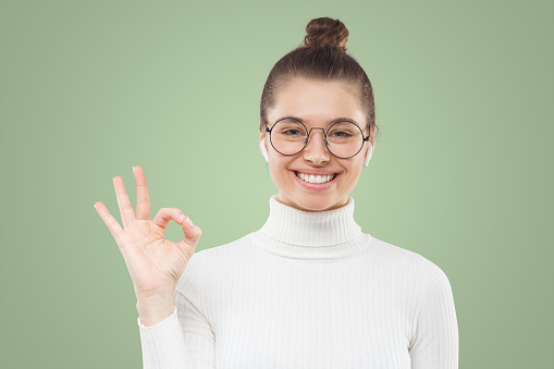 Approved! OK gesture. Young woman in glasses and white sweater showing okay sign, feelling confident about the quality of product, isolated on green background