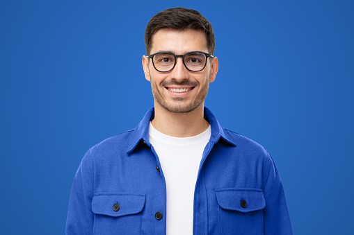 Close-up portrait of young handsome smiling man wearing blue shirt and glasses, isolated on blue background