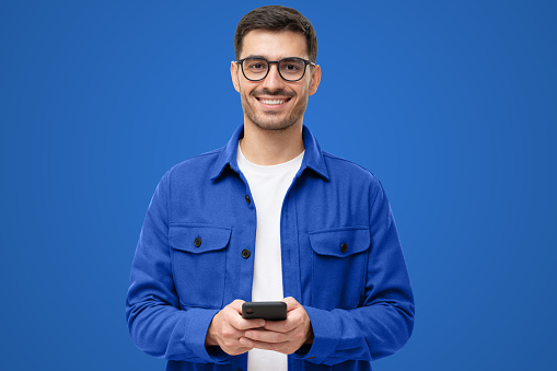 Young man in casual blue shirt and glasses, holding phone, looking at camera with smile, isolated on blue background